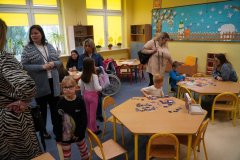Dzieci i dorośli w kolorowej sali świetlicy. Na pierwszym planie chłopiec układa puzzle przy sześciobocznym stoliku, a w tle widać inne dzieci bawiące się klockami oraz rodziców rozmawiających z opiekunami na tle zimowych dekoracji ściennych.