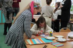 Dzień otwarty w szkole: dwie dziewczynki wspólnie pracują przy stoliku podczas warsztatów plastycznych. Jedna z nich smaruje papier klejem, a obok leżą kolorowe arkusze, wycięte kółka i flamastry. W tle widać innych uczestników wydarzenia.