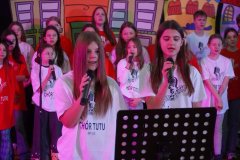 Występ chóru szkolnego podczas dnia otwartego. Grupa uczniów w czerwonych i białych koszulkach z logo „Chór Tutu” śpiewa na scenie. Na pierwszym planie dziewczyna śpiewa do mikrofonu, a w tle widać kolorowe dekoracje przedstawiające budynki.