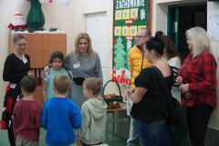 Dzień otwarty w szkole: grupa rodziców i dzieci rozmawia z nauczycielkami na korytarzu szkolnym. W tle widoczna jest gazetka ścienna o zachowaniu oraz świąteczne dekoracje, w tym figurka Mikołaja na szafce. Na stoliku obok stoi koszyk ze słodyczami.