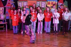 Dzień otwarty w szkole: występ artystyczny na scenie. Na pierwszym planie dziewczyna z niebieskimi włosami w ekspresyjnej pozie tanecznej. W tle stoi grupa uczniów z chóru szkolnego „Chór Tutu”, a za nimi widać kolorowe dekoracje przedstawiające bajkowe kamienice.