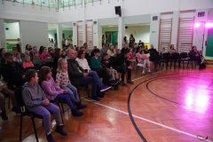 Dzień otwarty w szkole: liczna publiczność, składająca się z rodziców i dzieci, siedzi na krzesłach ustawionych w sali gimnastycznej. Widzowie z uwagą obserwują występy na oświetlonym różowym światłem parkiecie. W tle widać drabinki gimnastyczne oraz antresolę budynku.