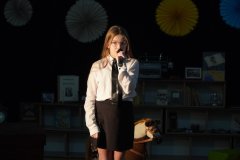 Dziewczynka w stylizacji przedwojennej stoi na scenie i mówi do mikrofonu.