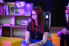 Na zdjęciu widać dziewczynkę z długimi włosami ozdobionymi czerwonymi pasemkami, ubraną w czarną koszulkę i niebieską tiulową spódnicę. Ma na sobie kolorowe bransoletki i naszyjnik oraz mikrofon na głowie. Siedzi na kolorowych siedziskach w oświetlonym fioletowym światłem pomieszczeniu, w tle widać półki z książkami.