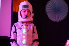 Uczeń w nowoczesnym stroju podobnym do stroju astronauty wypowiada swoją kwestię w przedstawieniu.