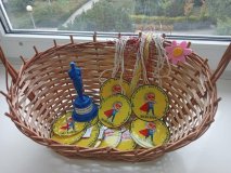 Na białym parapecie sali lekcyjnej, gdzie w tle znajduje się zakrzewione otoczenie szkoły znajduje się wiklinowy koszyczek. 'W nim jest mnóstwo własnoręcznie wykonanych żółtych medali z ilustarcją SuperChłopaka oraz napisem "Najfajniejszy Chłopak w klasie". Obok medali znajduje się również niebieska statuetka w formie mężczyzny z napisem "Najpopularniejszy Nauczyciel 2025/2026". 