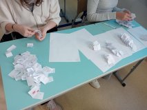 Ławka w sali lekcyjnej, a przy niej dwie dziewczynki liczące głosy w wyborach na Najpopularniejszego Nauczyciela w roku szkolnym 2025/2026. Przed nimi dużo kartek z nawiskami naczycieli odpowiednio posegregowanych.