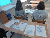 Ławka w sali lekcyjnej, a przy niej dwie dziewczynki liczące głosy w wyborach na Najpopularniejszego Nauczyciela w roku szkolnym 2025/2026. Za nimi ławka z dużą ilością kartek z nawiskami naczycieli (odpowiednio posegregowanych).