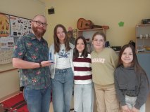 Sala lekcyjna- muzyczna- w tle gazetka dekoracyjna i  instrumenty muzyczne. Na pierwszym planie stoi "Najpopularniejszy Nauczyciel w roku szkolnym 2025/2026"- p. Łukasz Grocki, a w ręku trzyma niebieską statuetkę. Towarzyszą mu cztery przedstawicieli Samorządu Uczniowskiego. Wszyscy są w pogodnych nastrojach.