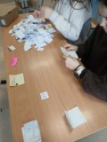 Uczniowie siedzą przy szkolnej ławce i liczą głosy oddane w tajnym głosowaniu. Na stole leży dużo złożonych karteczek z zapisanymi nazwiskami, które są segregowane i odczytywane podczas podliczania wyników akcji Samorządu Uczniowskiego z okazji Dnia Kobiet.