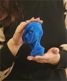 W rękach nauczycielki znajduje się wydrukowana na drukarce 3D niebieska statuetka przedstawiająca szyję i głowę kobiety w bujnej, fantazyjnej fryzurze. obrazek wyrozniajacy Na jasnoczerwonym, prążkowanym tle znajdują się ilustracje jasnych kwiatów, zdjęcia fioletowego medalu dla Najsymaptyczniejezej Dziewczyny oraz niebieskiej statuetki dla Najspympatyczniejszej Nauczycielki- oba w kształcie kwiatka, a także napis "SZKOLNE OBCHODY Dnia Kobiet".