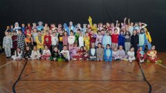 Uczniowie SP 22 w Płocku podczas Dnia Piżamy – wspólne zdjęcie w sali gimnastycznej, na tle zielonej kotary. Dzieci pozują w kolorowych piżamach, kombinezonach, szlafrokach i innych wygodnych strojach domowych, trzymając poduszki i pluszowe zabawki. 