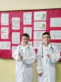 Uczniowie w strojach lekarskich pozują z  receptami zalecającymi spacer na świeżym powietrzu na tle gazetki tematycznej