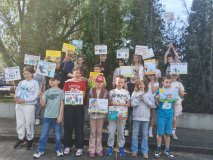 Na zdjęciu widać Dzieci przebywające na szkolnym terenie, wśród drzew i krzewów. Uczniowie trzymają własnoręcznie wykonane plakaty z rysunkami i hasłami dotyczącymi ochrony środowiska- sprzątania śmieci, sprzątania po psach... 