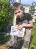 Na zdjęciu widać Chłopca znajdującego się na terenie szkolnego skweru. Jest On lekko pochylony i trzyma w ręku drewniany patyk z przymocowanym plakatem. Na kartce znajduje się obrazek psa oraz napis: ,,Twój pies- sprzątaj po swoim psie”. 