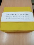 Żółte pudełko stojące na drewnianym blacie. Na pudełku znajduje się biała kartka z napisem „Skrzynka Życzliwości – 21 listopada – Dzień Życzliwości i Pozdrowień. Wrzuć dla kogoś miłą kartkę, dobre słowo lub pochwałę! (18.11–20.11 do 14:00) 