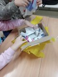 Dwie osoby otwierają pudełko, z którego wystaje mnóstwo złożonych karteczek z życzeniami i pozdrowieniami. Na zdjęciu widoczne są ręce uczniów przeglądających kolorowe i białe kartki. Pudełko ma rozerwane, żółte oklejenie. 
