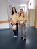 Dwie uczennice w jasnożółtych koszulkach stoją na szkolnym korytarzu. Każda trzyma wiklinowy koszyk z kopertami i karteczkami. Korytarz jest jasny, z żółtymi szafkami po prawej stronie. 