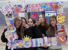 Grupa uczennic trzymają kolorową ramkę ozdobioną naklejkami i napisami w tematyce Dnia Kobiet. Wszystkie uśmiechają się, pozując do zdjęcia na tle szkolnej dekoracji na Dzień Kobiet w SP 22 Płock.