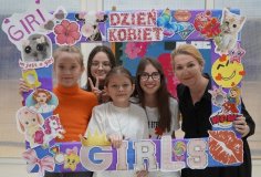 Grupa uczennic wraz z nauczycielką trzymają kolorową ramkę ozdobioną naklejkami i napisami w tematyce Dnia Kobiet. Wszystkie uśmiechają się, pozując do zdjęcia na tle szkolnej dekoracji na Dzień Kobiet w SP 22 Płock.