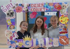 Trzy uczennice trzymają kolorową ramkę ozdobioną naklejkami i napisami w tematyce Dnia Kobiet. Wszystkie uśmiechają się, pozując do zdjęcia na tle szkolnej dekoracji na Dzień Kobiet w SP 22 Płock.