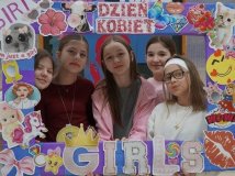 Grupa uczennic trzymają kolorową ramkę ozdobioną naklejkami i napisami w tematyce Dnia Kobiet. Wszystkie uśmiechają się, pozując do zdjęcia na tle szkolnej dekoracji na Dzień Kobiet w SP 22 Płock.