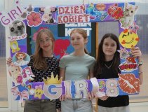 Trzy uczennice trzymają kolorową ramkę ozdobioną naklejkami i napisami w tematyce Dnia Kobiet. Wszystkie uśmiechają się, pozując do zdjęcia na tle szkolnej dekoracji na Dzień Kobiet w SP 22 Płock