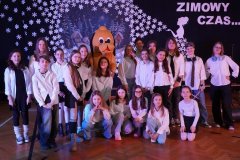 Grupa dzieci i młodzieży w białych koszulach i czarnych spodniach lub spódnicach pozuje na tle zimowej dekoracji z napisem „ZIMOWY CZAS…”. W centrum stoi osoba w kostiumie maskotki przypominającej psa. Scena udekorowana jest śnieżynkami i obrazem zimowego krajobrazu.
