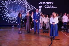 Kobieta w białej bluzce i granatowej spódnicy przemawia do mikrofonu na tle zimowej dekoracji z napisem „ZIMOWY CZAS…”. Obok niej stoją trzy osoby w eleganckich strojach, a w tle znajduje się grupa uczniów ubranych na biało, przygotowanych do występu.