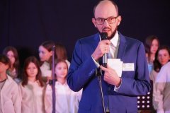 Mężczyzna w okularach, ubrany w niebieski garnitur i białą koszulę, przemawia do mikrofonu. W tle widoczne są dzieci ubrane w jasne stroje, stojące w szeregu na scenie.