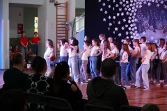 Grupa dzieci w białych koszulkach i dżinsach występuje na scenie przed publicznością. W tle dekoracja zimowa z domkami i śnieżynkami. Po lewej stronie stoją dziewczynki w czerwonych koszulkach, obserwujące występ.
