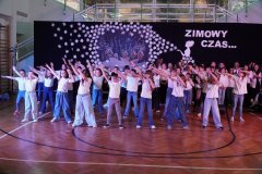 Grupa dzieci w białych koszulkach i dżinsach występuje na scenie przed publicznością. W tle dekoracja zimowa z domkami i śnieżynkami. Po lewej stronie stoją dziewczynki w czerwonych koszulkach, obserwujące występ.