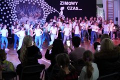 Grupa dzieci w białych koszulkach i dżinsach występuje na scenie przed publicznością. W tle dekoracja zimowa z domkami i śnieżynkami. Po lewej stronie stoją dziewczynki w czerwonych koszulkach, obserwujące występ.