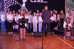 Grupa dzieci ubranych na biało i czarno występuje na scenie, a jedna dziewczynka na pierwszym planie trzyma mikrofon i śpiewa.