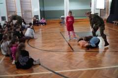 Dwie Dziewczynki demonstrują, jak należy udzielić pierwszej pomocy – jedna klęczy i próbuje nawiązać kontakt z poszkodowanym, druga natomiast czeka na polecenia i wzywa pomoc specjalistyczną. Po lewej stronie trwa rozmowa drugiej Prowadzącej z grupą Chłopców, którzy zadają pytania związane z tematem szkolenia.