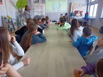 czytelnia biblioteki, kobieta prowadzi zajęcia, przy długim stole siedzi grupa dzieci i słucha pogadanki, w tle baner z napisem „Bądźmy eko z biblioteką”