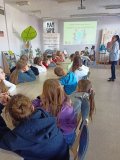 czytelnia biblioteki, kobieta prowadzi pogadankę, przy długim stole siedzi grupa dzieci i słucha, w tle baner z napisem „Projekt edukacyjny dla klas 3. Bądźmy eko z biblioteką”
