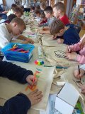długi stół, przy którym siedzi grupa uczniów zajęta wykonywaniem flamastrami rysunków na ułożonych na blacie torbach bawełnianych