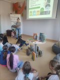 Grupa dzieci siedzi na podłodze i z uwagą słucha edukatorki z Przedsiębiorstwa Gospodarowania Odpadami w Płocku.