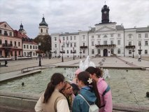 Grupa dzieci rozwiązuje zadanie przy fontannie na Starym Rynku. W tle widoczny ratusz.