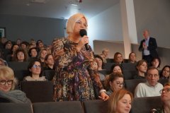 Kobieta stojąca z mikrofonem. Wokół niej kobiety i mężczyźni siedzący w auli widowiskowej.