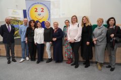 Grupa 9 kobiet i 2 mężczyzn stojących w linii przed sceną. Za nimi w tle stoi kolorowy plakat z napisami.