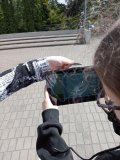 foto_3 Zdjęcie w plenerze- plac przed szkołą: jedna z uczennic trzyma w ręku tablet z uruchomionym czujnikiem promieniowania UV, a druga trzyma bezpośrednio nad czujnikiem szkło z okularów przeciwsłonecznych