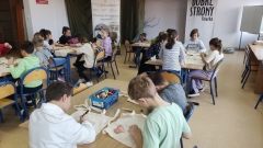 Dzieci, siedząc przy stole, rysują kredkami i malują farbami bawełniane torby na zakupy.