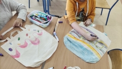 Dzieci, siedząc przy stole, rysują kredkami i malują farbami bawełniane torby na zakupy.