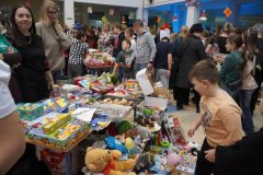 Gwarny kiermasz świąteczny w szkole lub świetlicy. Stoły wypełnione kolorowymi zabawkami, grami planszowymi i domowymi wypiekami. Wokół stoisk tłoczą się dorośli i dzieci, a w tle widać świąteczne dekoracje.