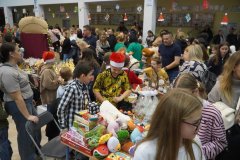 Gwarny kiermasz świąteczny w szkole lub świetlicy. Stoły wypełnione kolorowymi zabawkami, grami planszowymi i domowymi wypiekami. Wokół stoisk tłoczą się dorośli i dzieci, a w tle widać świąteczne dekoracje.