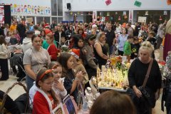Gwarny kiermasz świąteczny w szkole lub świetlicy. Stoły wypełnione kolorowymi zabawkami, grami planszowymi i domowymi wypiekami. Wokół stoisk tłoczą się dorośli i dzieci, a w tle widać świąteczne dekoracje.