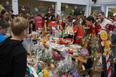 Gwarny kiermasz świąteczny w szkole lub świetlicy. Stoły wypełnione kolorowymi zabawkami, grami planszowymi i domowymi wypiekami. Wokół stoisk tłoczą się dorośli i dzieci, a w tle widać świąteczne dekoracje.
