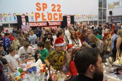 Tłum uczestników „Mikołajkowego Kiermaszu Rozmaitości” na forum Szkoły Podstawowej nr 22 w Płocku. W tle widnieje duży napis „SP 22 PŁOCK”, a na pierwszym planie dzieci i dorośli przeglądają stoiska z zabawkami, rękodziełem i biżuterią.